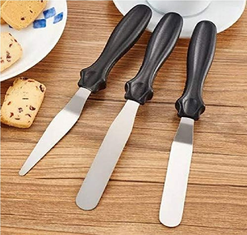 Three metal spatulas with black handles on a wooden surface next to cookies.